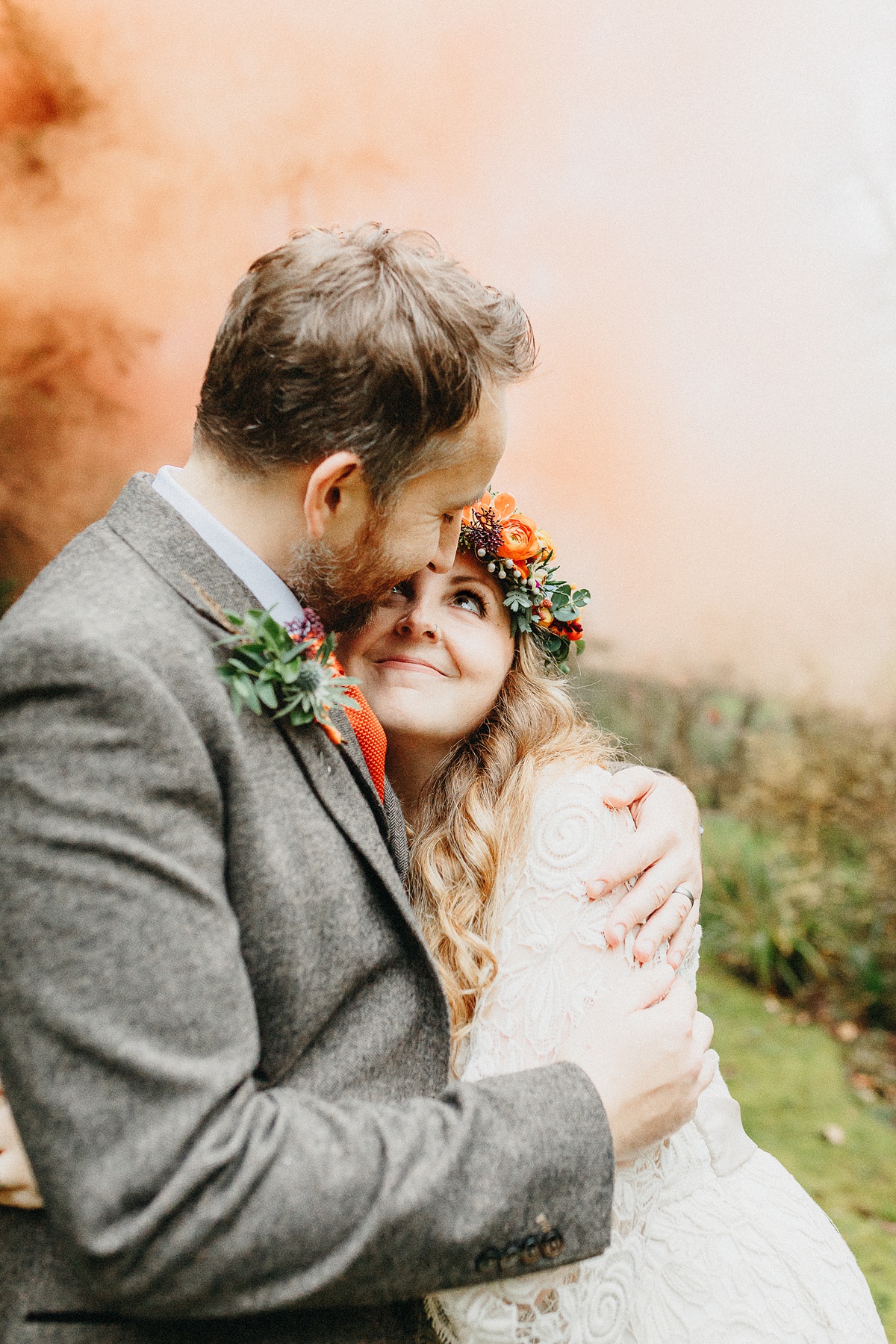 Bride and groom snuggle up whilst lookng at each other. Bride wear an orange and green flower crown. There is orange smoke in the background