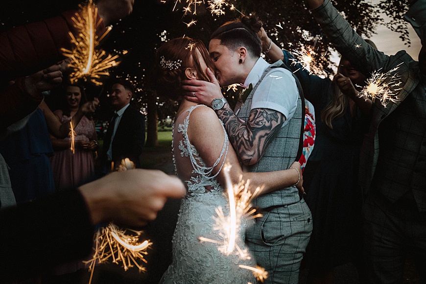 Bride and bride embrace and kiss as wedding guests waive sparklers around them