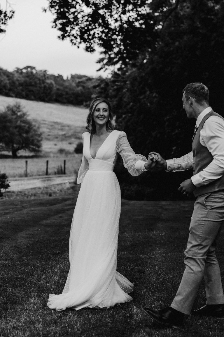 Bride and groom hold hands while smiling at each other and walking through the gardens of The Courtyard Venue Oswestry . Bride wears a Sassi Holford wedding dress.