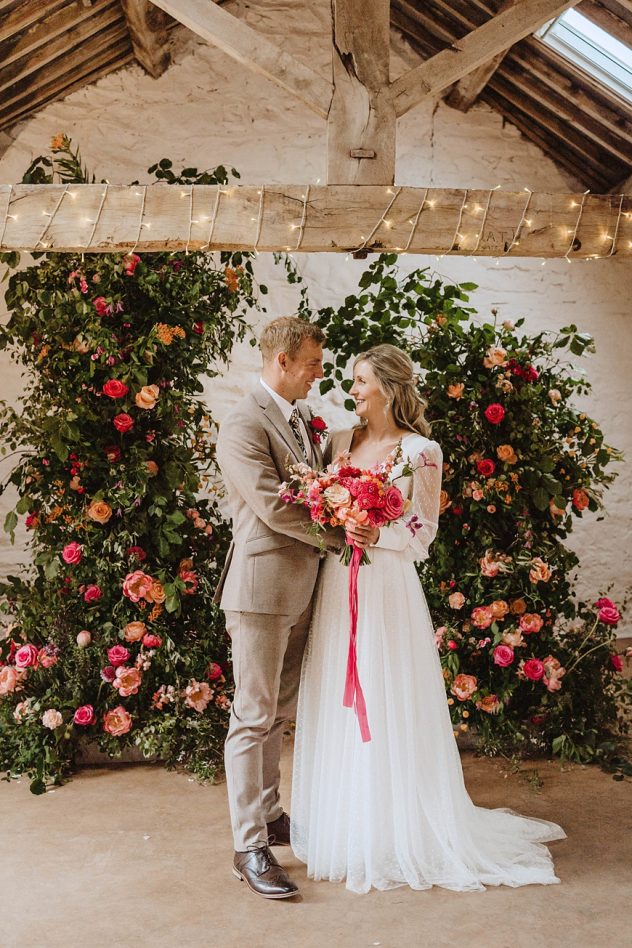 Bride and Groom and The Courtyard Venue stood in front of a broken flower arch with lots of foliage and hot pink flowers