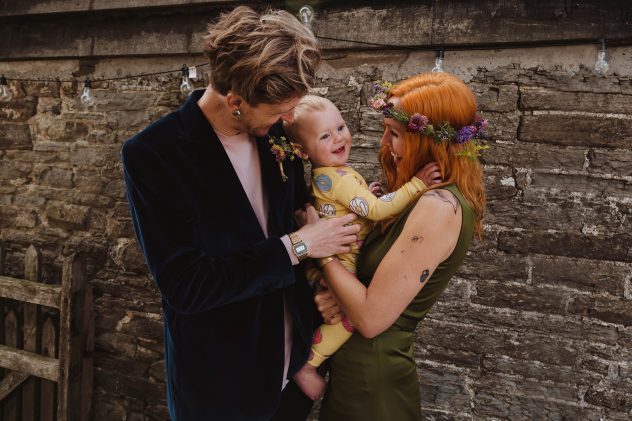 Family of three stood in front of wall at Wilde Lodge wedding. Mum wears a green gown and flower crown. Dad wears a blue velvet jacket. Baby wears a yellow print with donughts