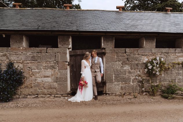 A wide shot of a couple leaning against a stable door at The Courtyard Venue. The bride wears a white gown and holds a pink bouquet. The Groom leans back against the door. He is wearing a stone coloured waistcoat and matching trousers