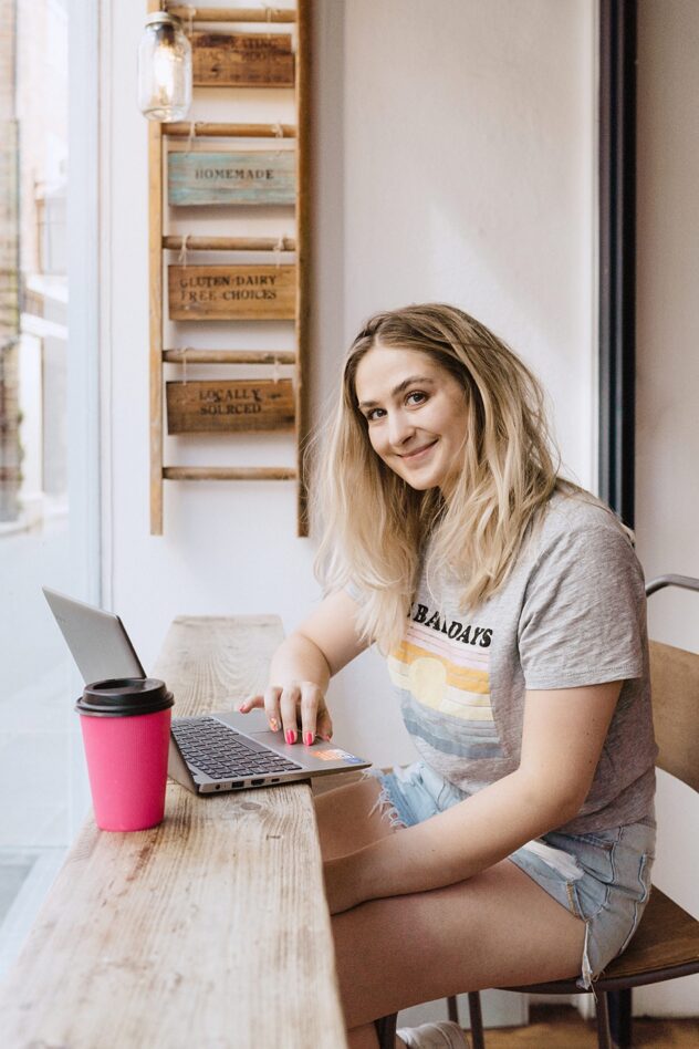 Woman smiling at camera whilst sat in coffee shop working on laptop. Brand storytelling
