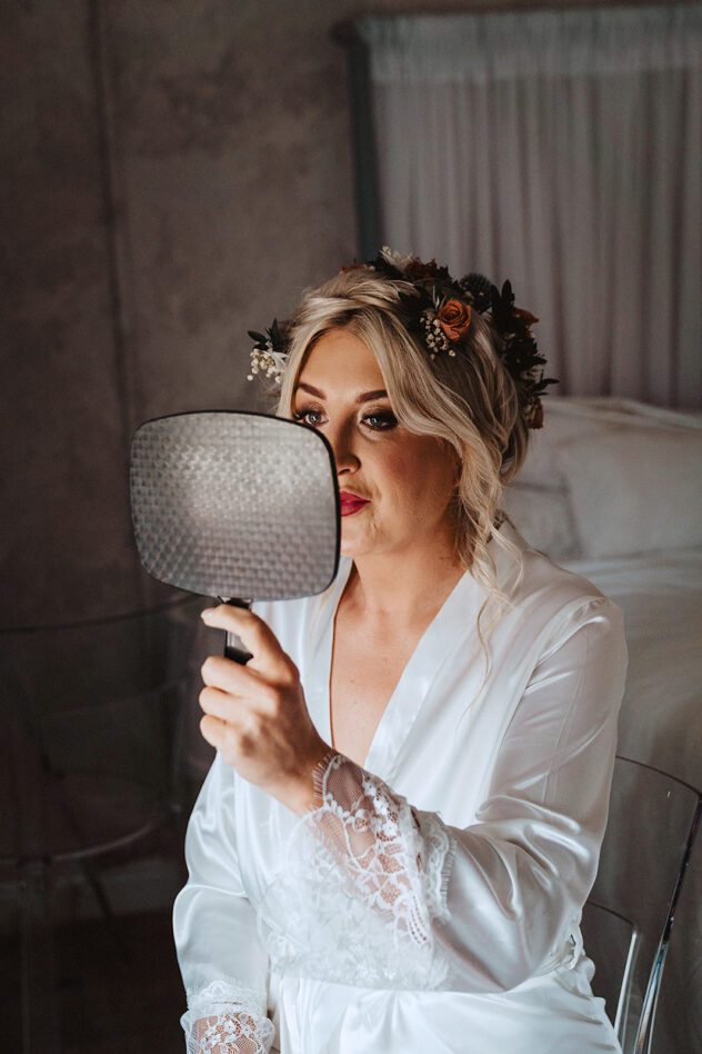 Bride holds mirror to her face, she is wearing a flower crown. Stanford farm wedding